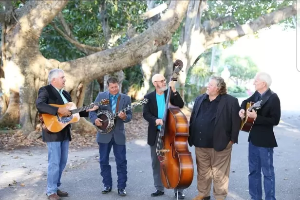 five older men in suit coats and jeans standing in front of a banyan tree holding a guitar, a banjo, an upright bass and another guitar.
